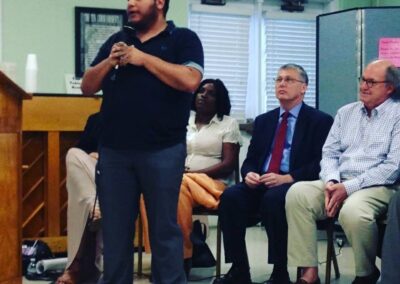 Jack Knoxville is a brown skinned trans man seen wearing a black polo style shirt with jeans and sneakers while holding a microphone during a campaign event in Knoxville, TN