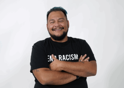 Jack is a medium brown skinned man with short black hair seen smiling and standing with his arms folded wearing a black shirt with white writing that says "end racism".